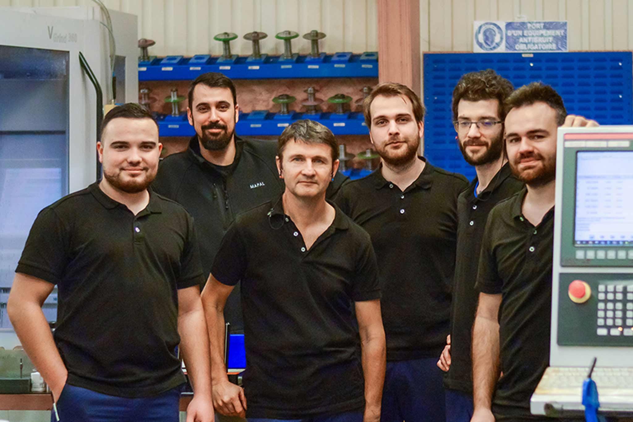 Eine Gruppe von sechs Männern mit schwarzen T-Shirts sind um eine Werkzeugmaschine gruppiert. 