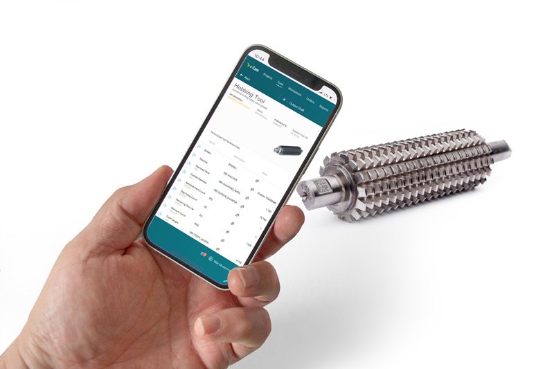 A hand shows the c-Com application MAS on a mobile phone. A hobbing tool can be seen in the background.